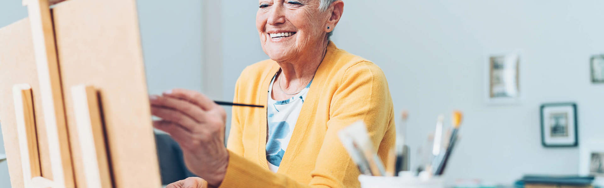 An older woman wearing a yellow cardigan sat at a table painting using a table top easel