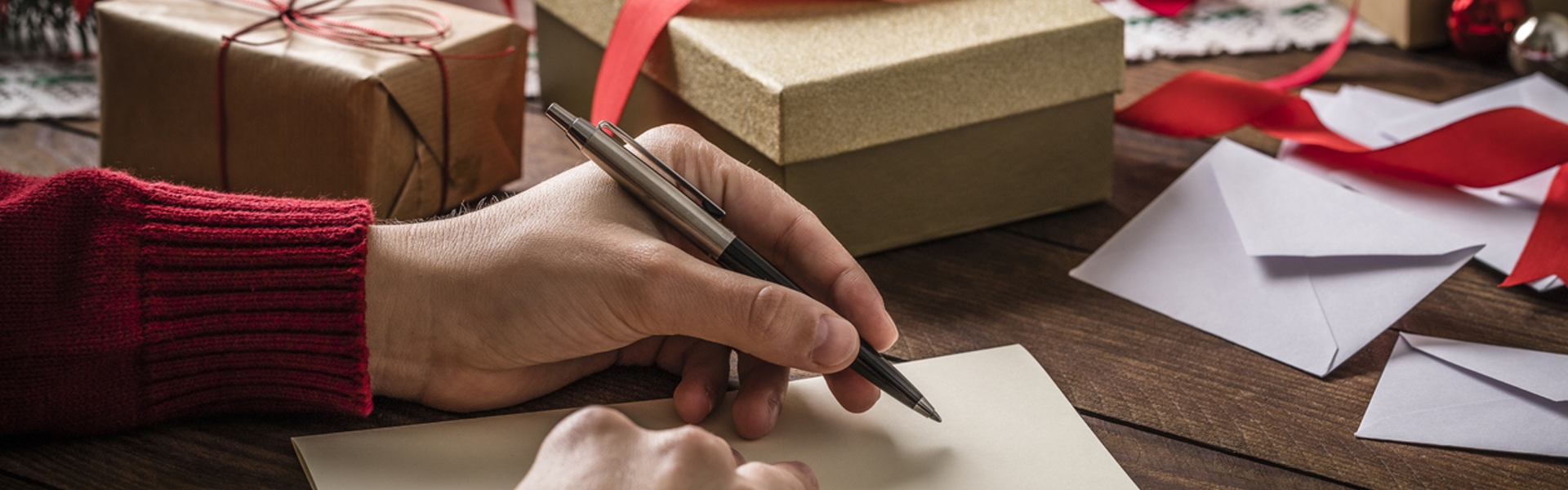 a pair of hands in a red jump writing a letter surrounded by brown Christmas parcels with red ribbons