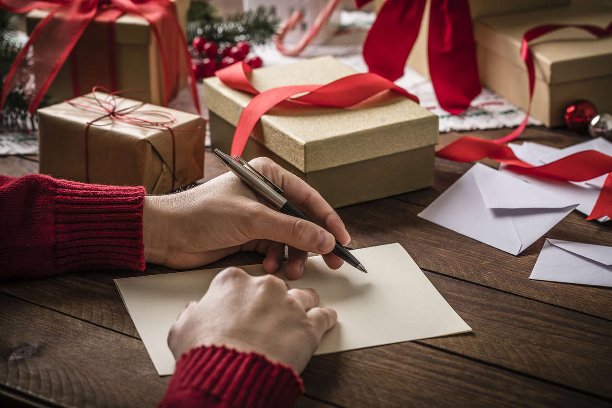 a pair of hands in a red jump writing a letter surrounded by brown Christmas parcels with red ribbons