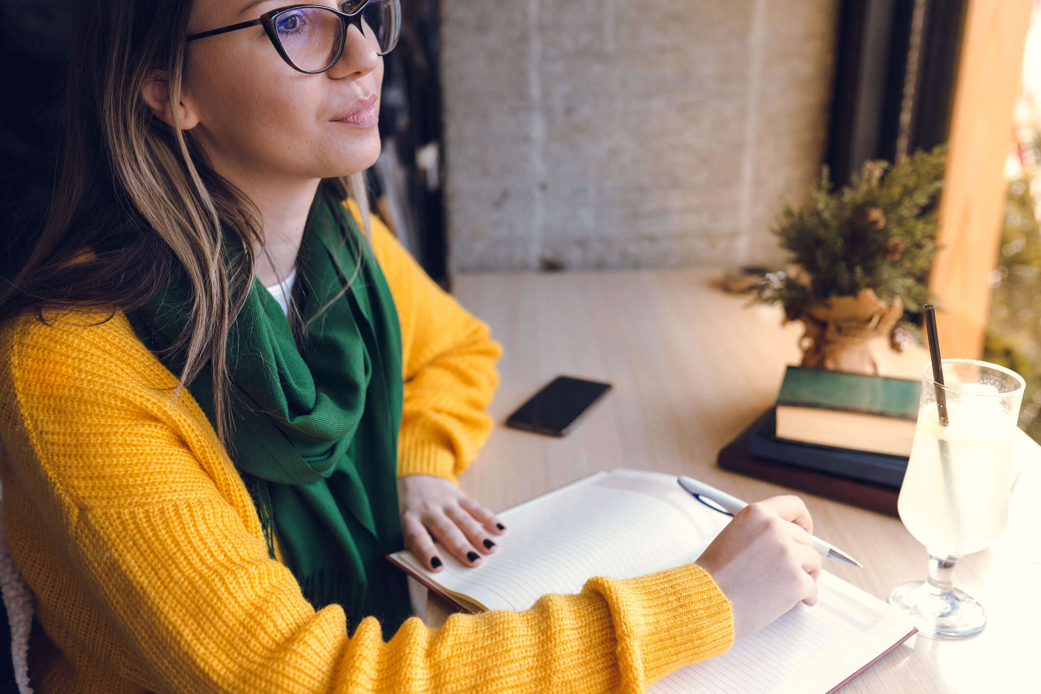 a woman wearing a yellow jumper and green scarf writing in a journal at a desk and looking out of the window