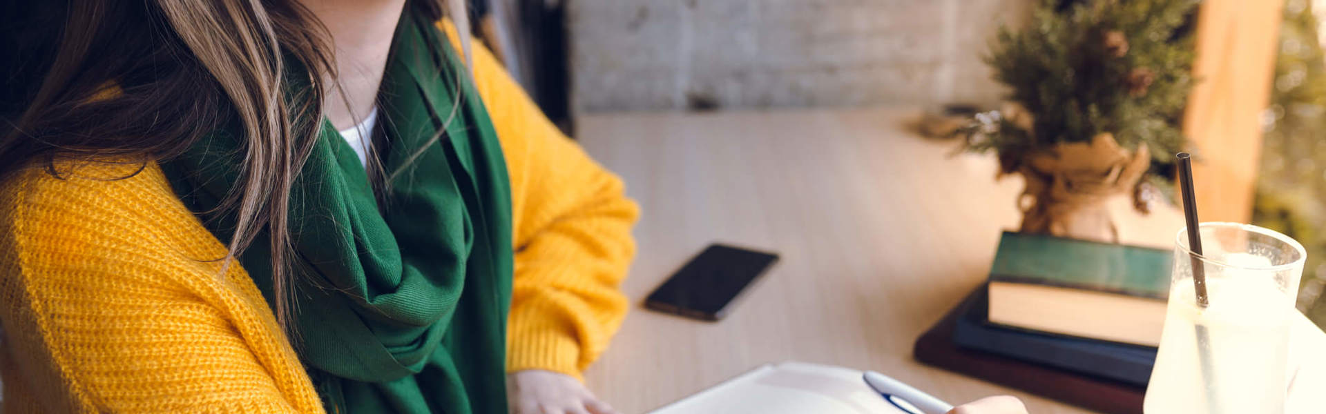 a woman wearing a yellow jumper and green scarf writing in a journal at a desk and looking out of the window