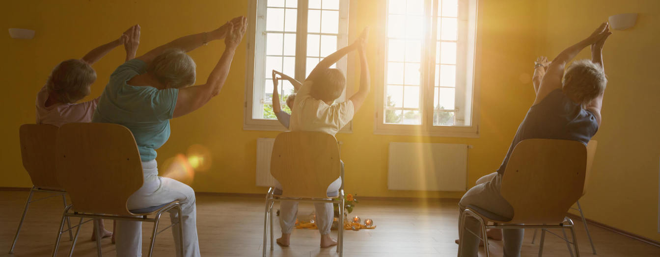 A group taking part in a seated exercise class, Sit Fit, with sun shining through the windows.
