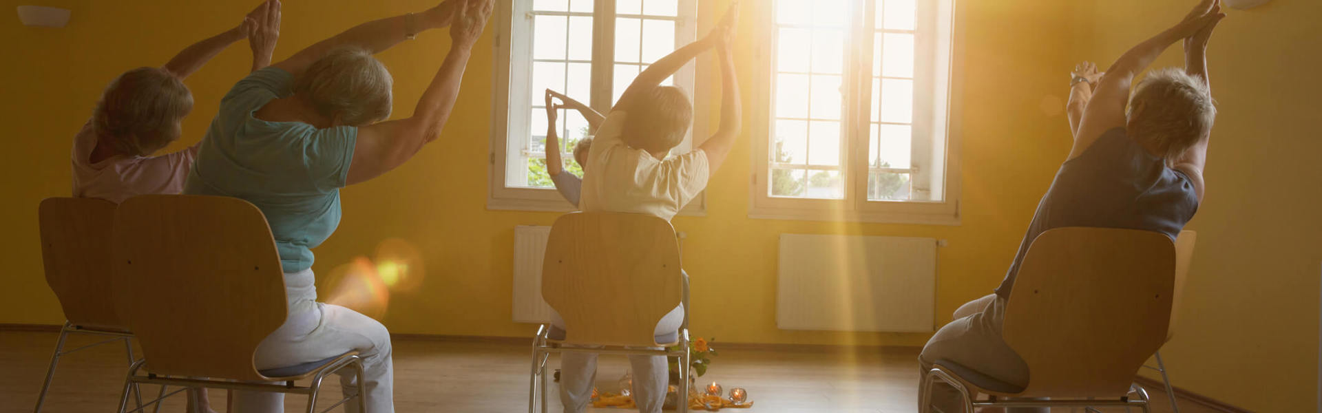 A group taking part in  a seated exercise class, Sit Fit, with sun shining through the windows.