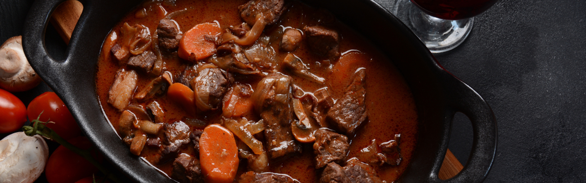 Main Image:  Beef stew in a black cast iron pan with vegetables and a glass of red wine