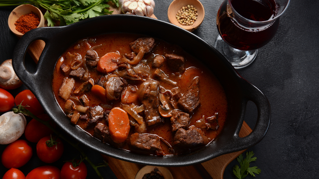 Main Image:  Beef stew in a black cast iron pan with vegetables and a glass of red wine