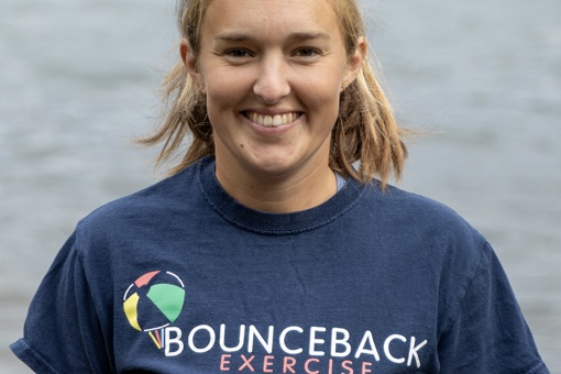 Headshot photo of Beth Boxall, Clinical Exercise Specialist with Bounceback Exercise, smiling wearing a navy blue t-shirt that reads "BOUNCEBACK EXERCISE with a beachball logo" in front of a body of water.