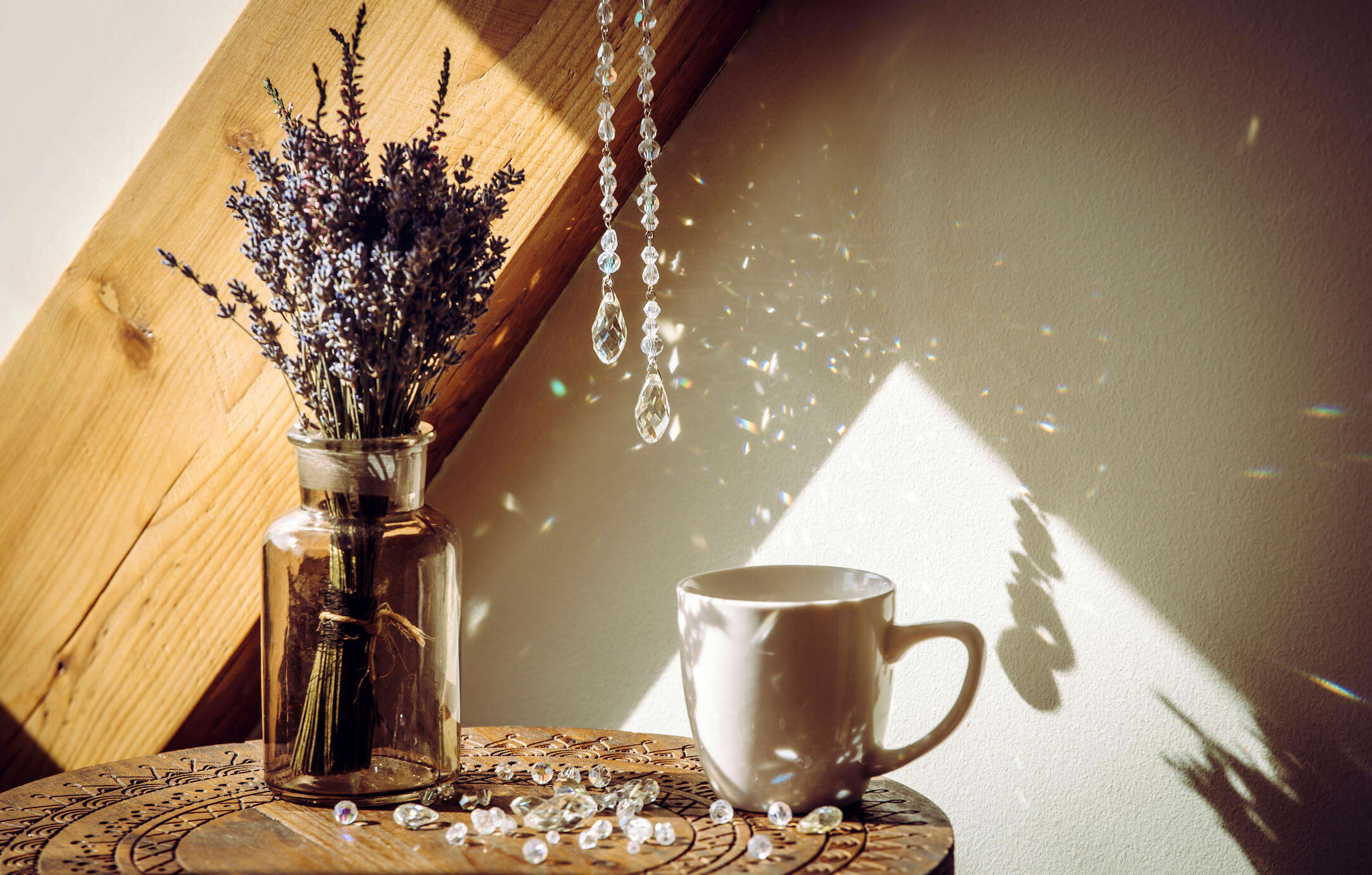 A jewelled sun catch hanging over a vase containing lavender and a white mug on a wooden table