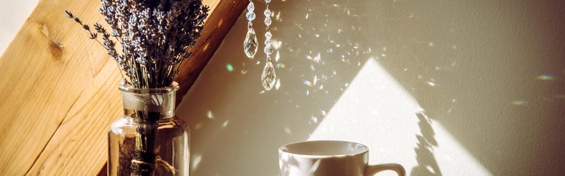 A jewelled sun catch hanging over a vase containing lavender and a white mug on a wooden table
