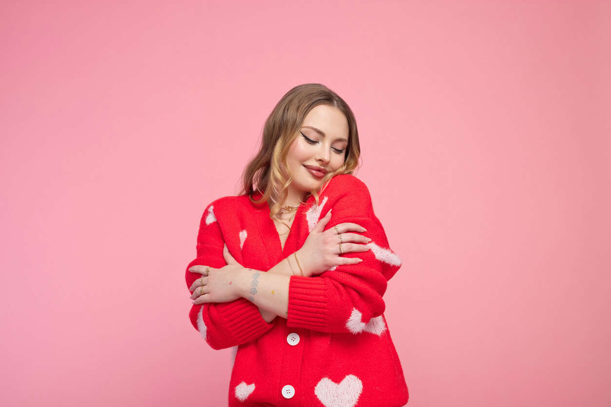 Valentine's portrait of a lovely young woman in a red heart cardigan, demonstrating self-love, set against a vibrant pink backdrop.