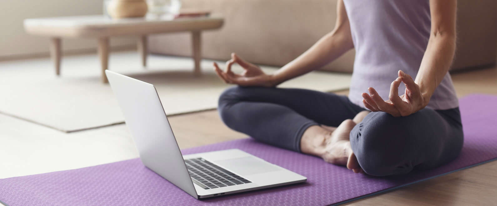 a person wearing a purple t-shirt sat cross legged on a  purple yoga mat doing yoga from a laptop