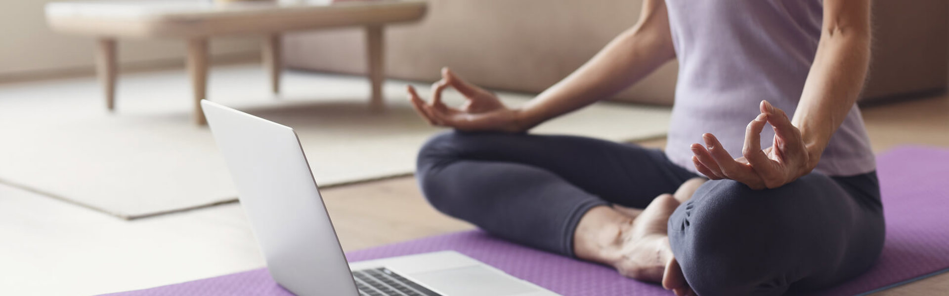 a person wearing a purple t-shirt sat cross legged on a purple yoga mat doing yoga from a laptop