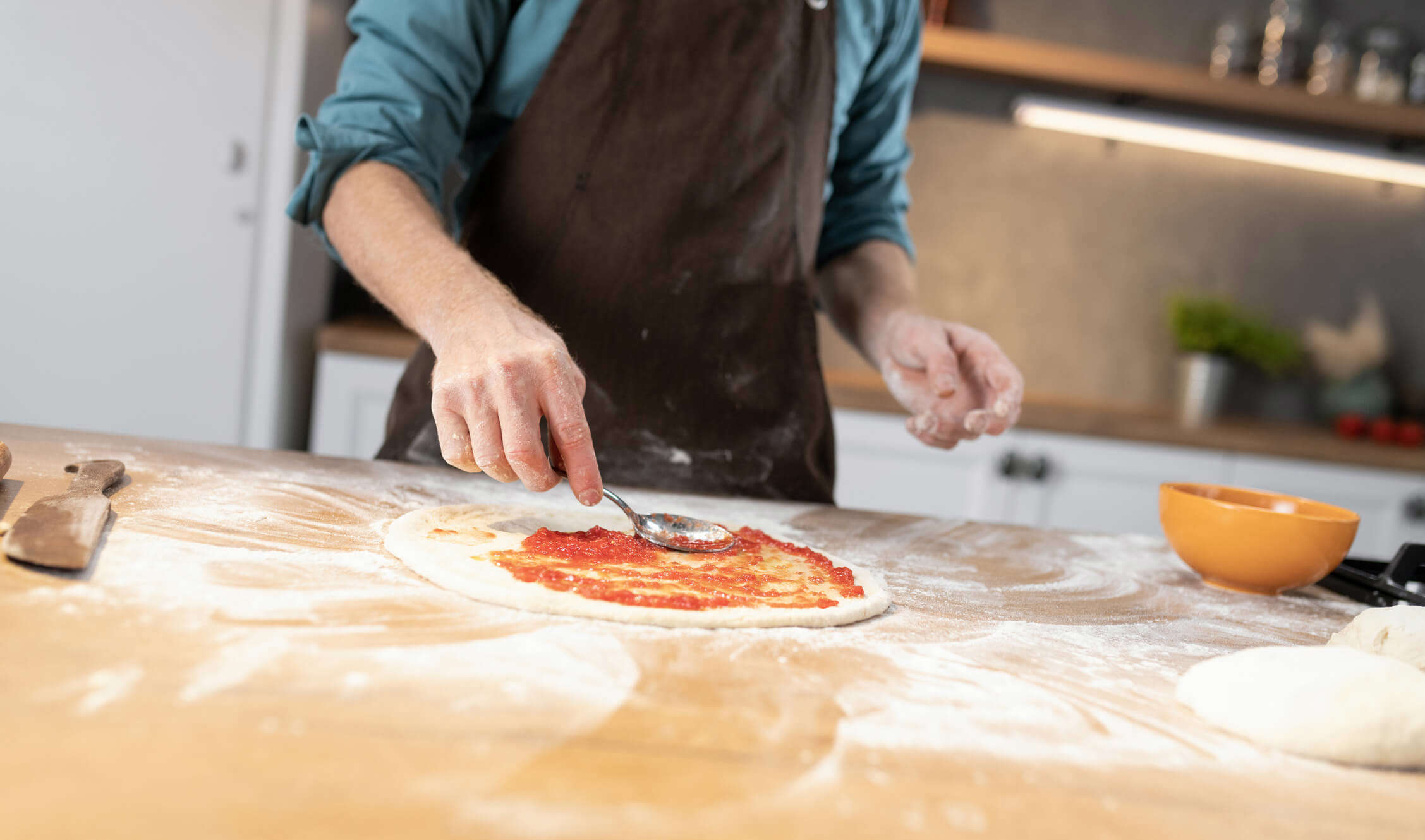someone spreading tomato passata on a pizza dough base on a floured surface with a spoon