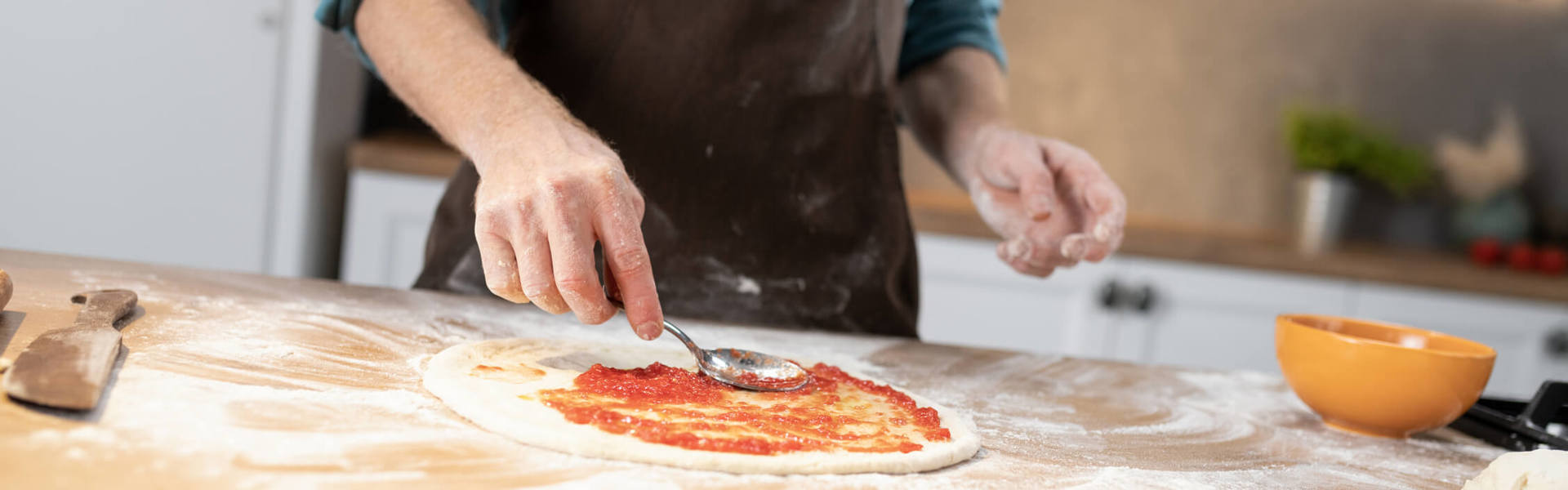 someone spreading tomato passata on a pizza dough base on a floured surface with a spoon