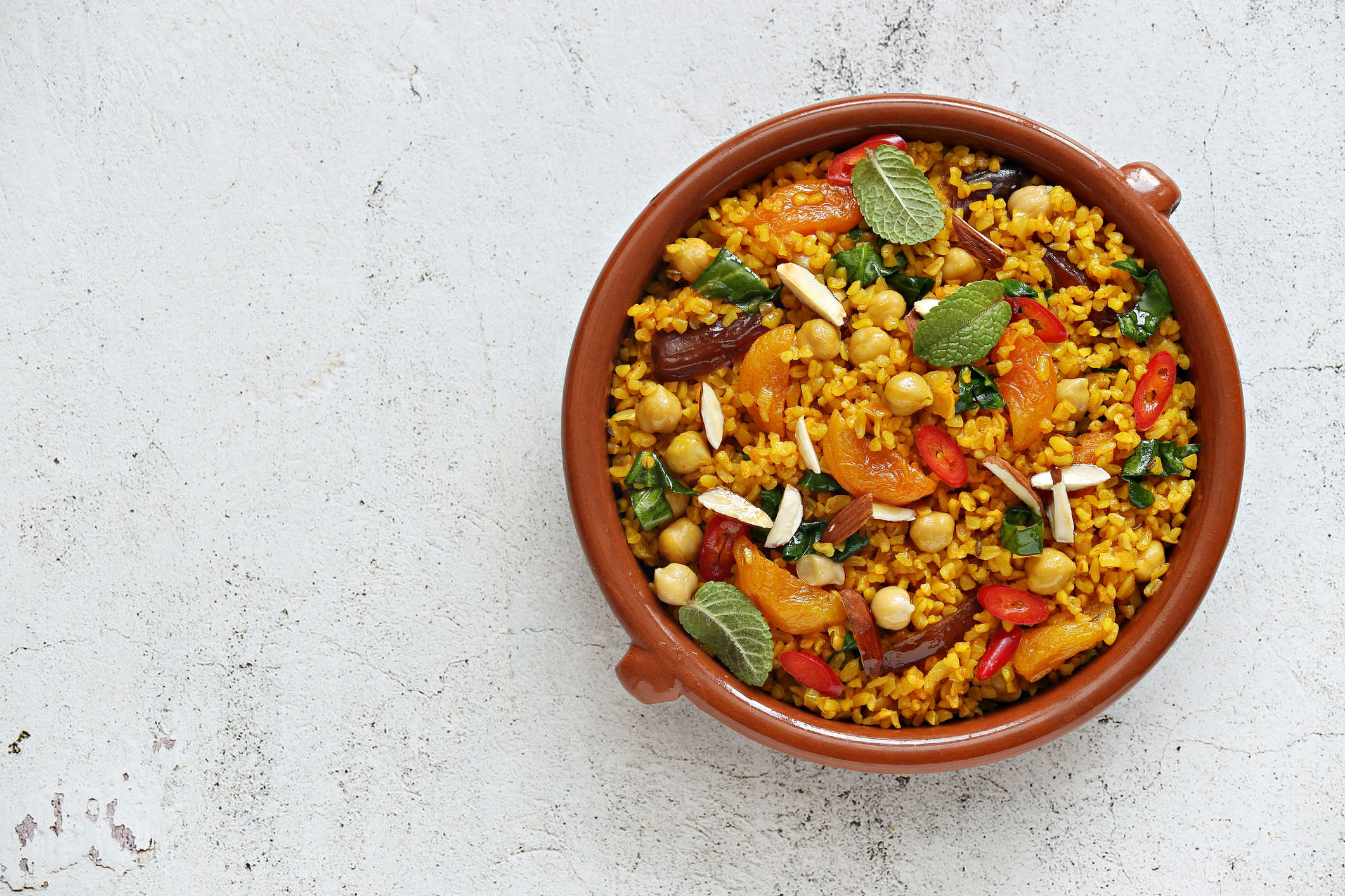 Main image: a bowl of Moroccan Steamed Couscous And Apricot Salad, a healthy vegan recipe, in a brown bowl on a while stone table top.