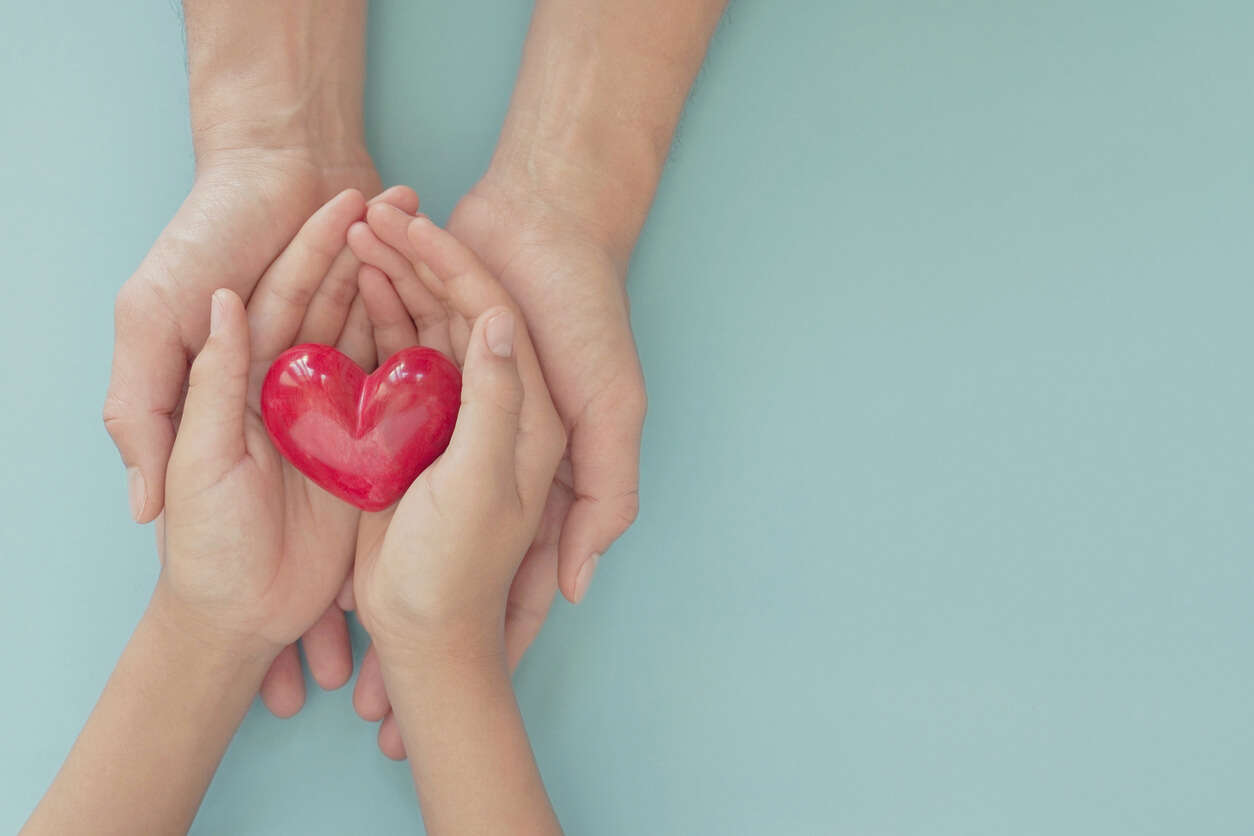 a pair of children's hands holding a red heart, being held by a pair of adult hands on a blue background