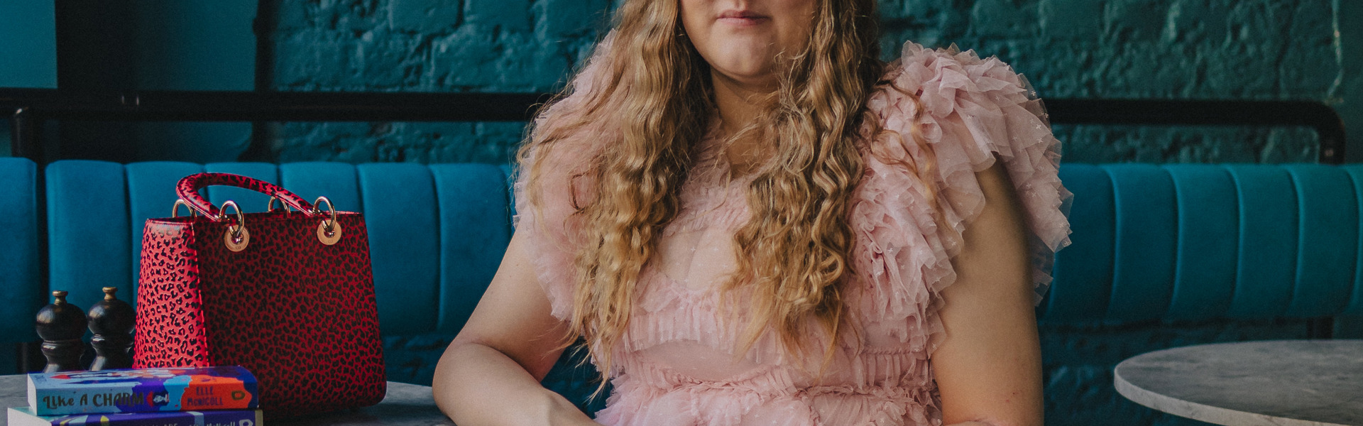 Main image: author Elle McNicoll in a frilly, floor length, light pink dress, sat in front of a dark turquoise wall in bar or café. 