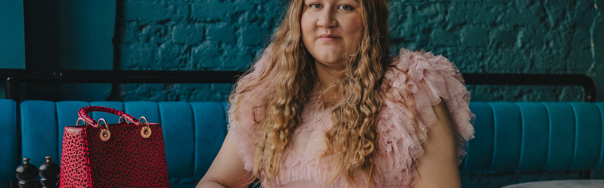 Main image: author Elle McNicoll in a frilly, floor length, light pink dress, sat in front of a dark turquoise wall in bar or café. 