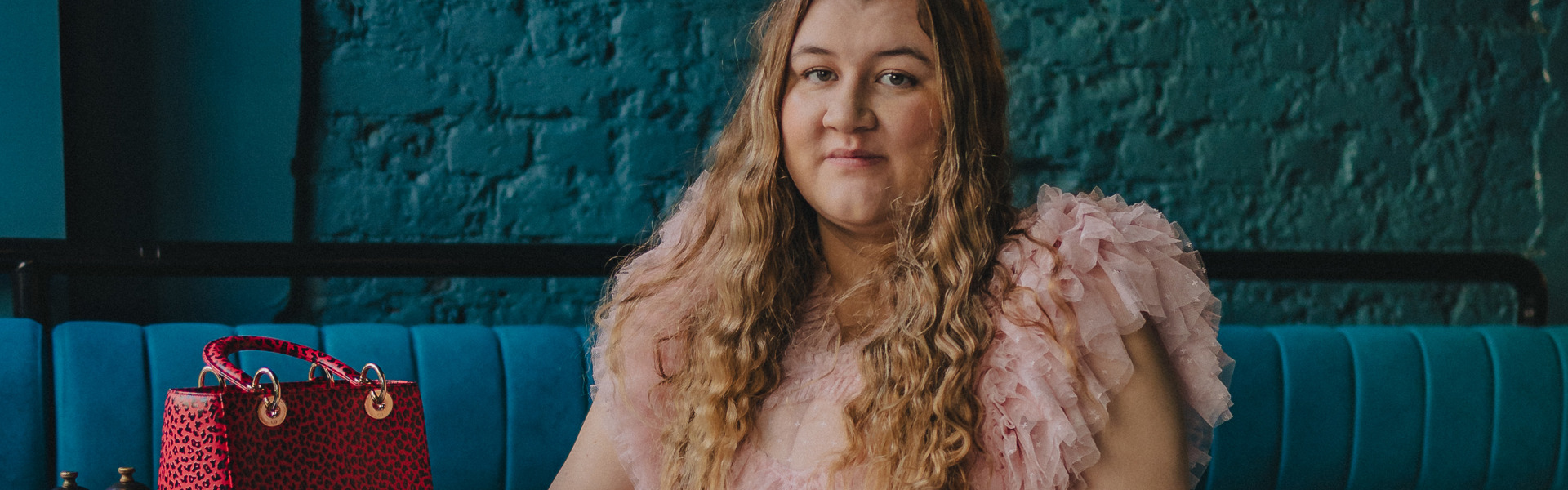 Main image: author Elle McNicoll in a frilly, floor length, light pink dress, sat in front of a dark turquoise wall in bar or café. 