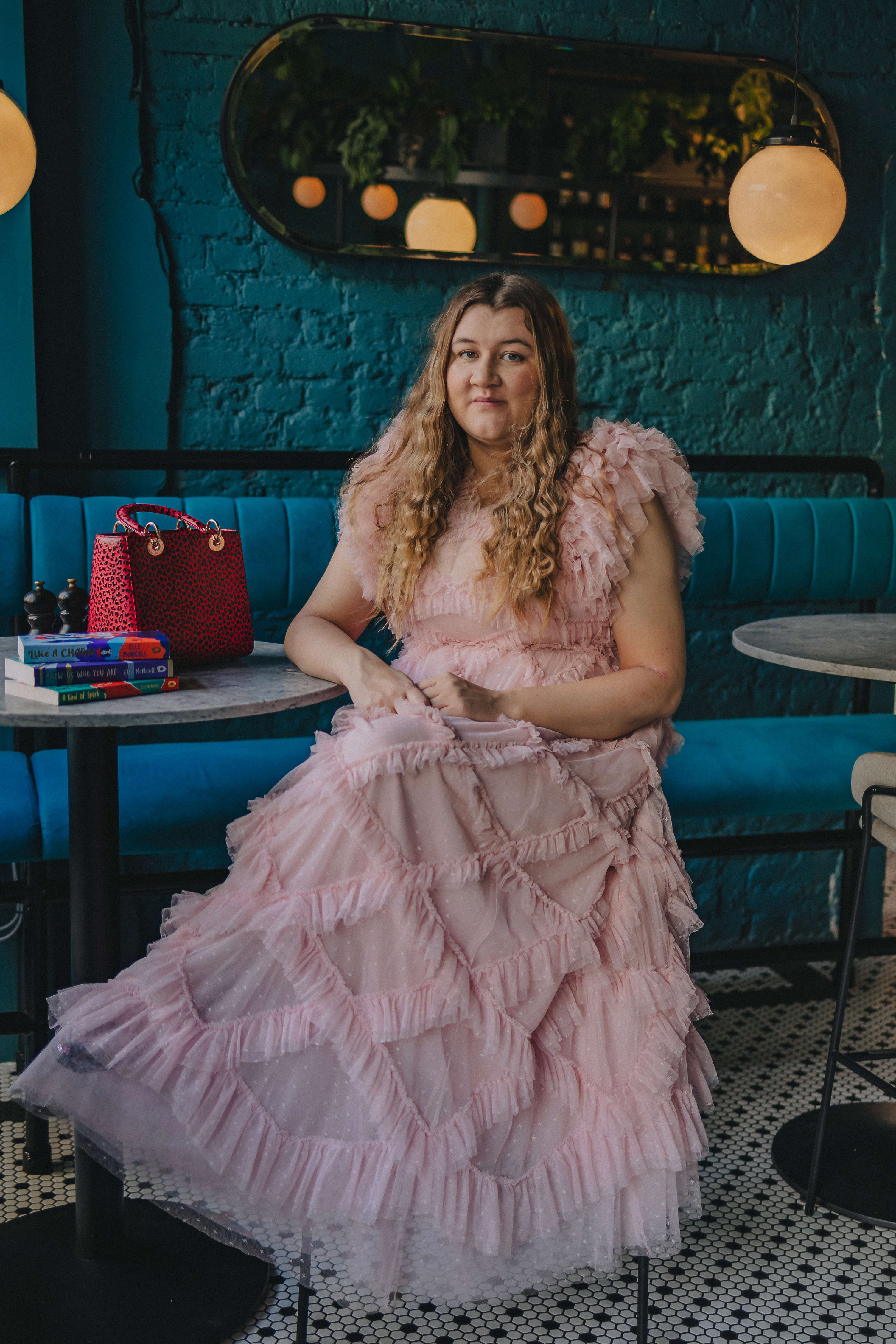Main image: author Elle McNicoll in a frilly, floor length, light pink dress, sat in front of a dark turquoise wall in bar or café. 
