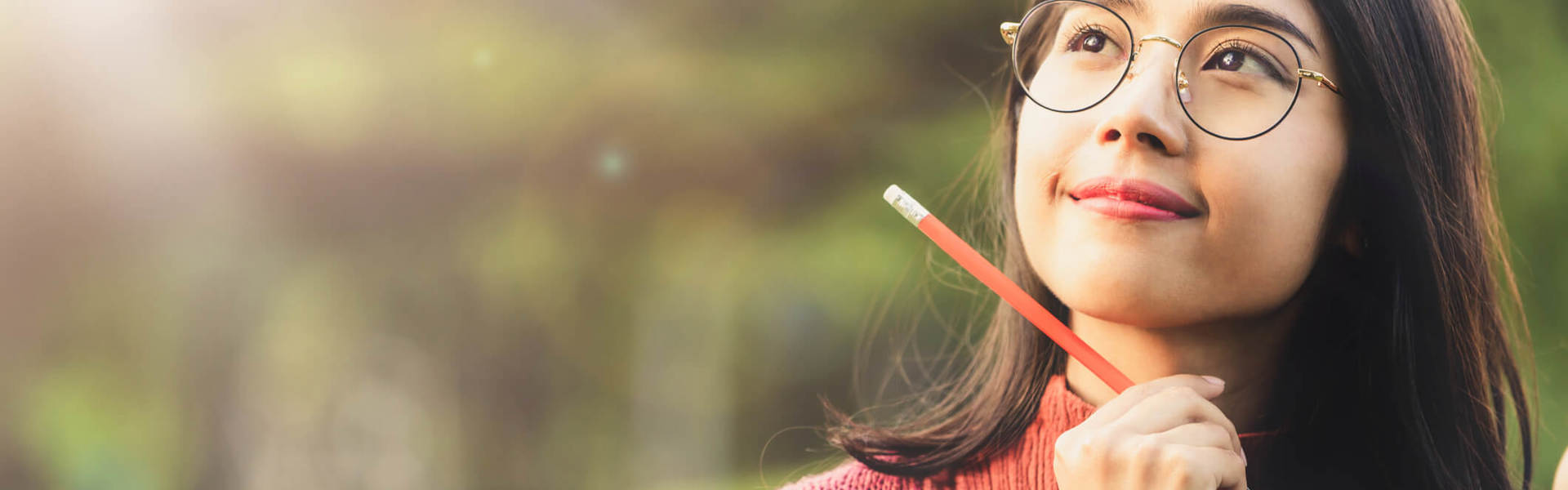 a woman wearing glasses and holding a pencil whilst resting her chin on an open green book looking up and thinking