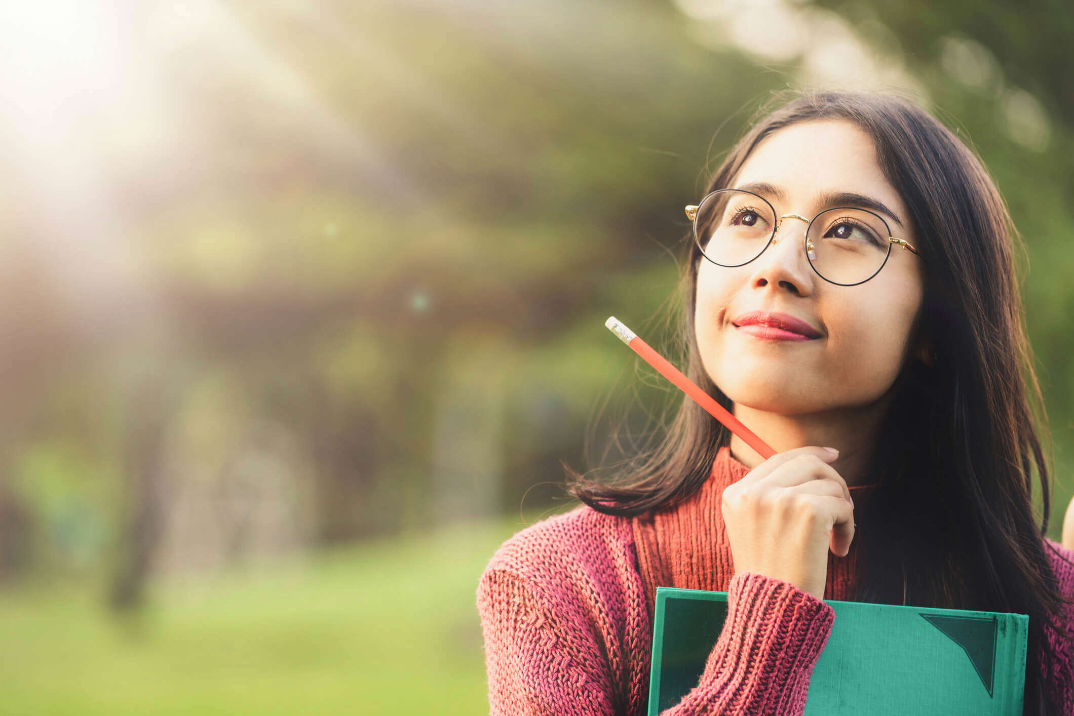 a woman wearing glasses and holding a pencil whilst resting her chin on an open green book looking up and thinking