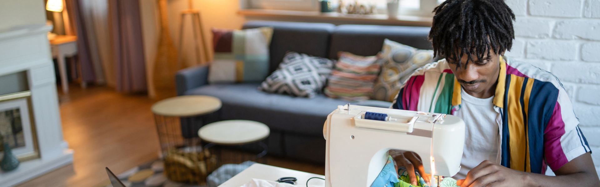 a young man wearing a colourful stripe short sleeved shirt sat at a sewing machine sewing