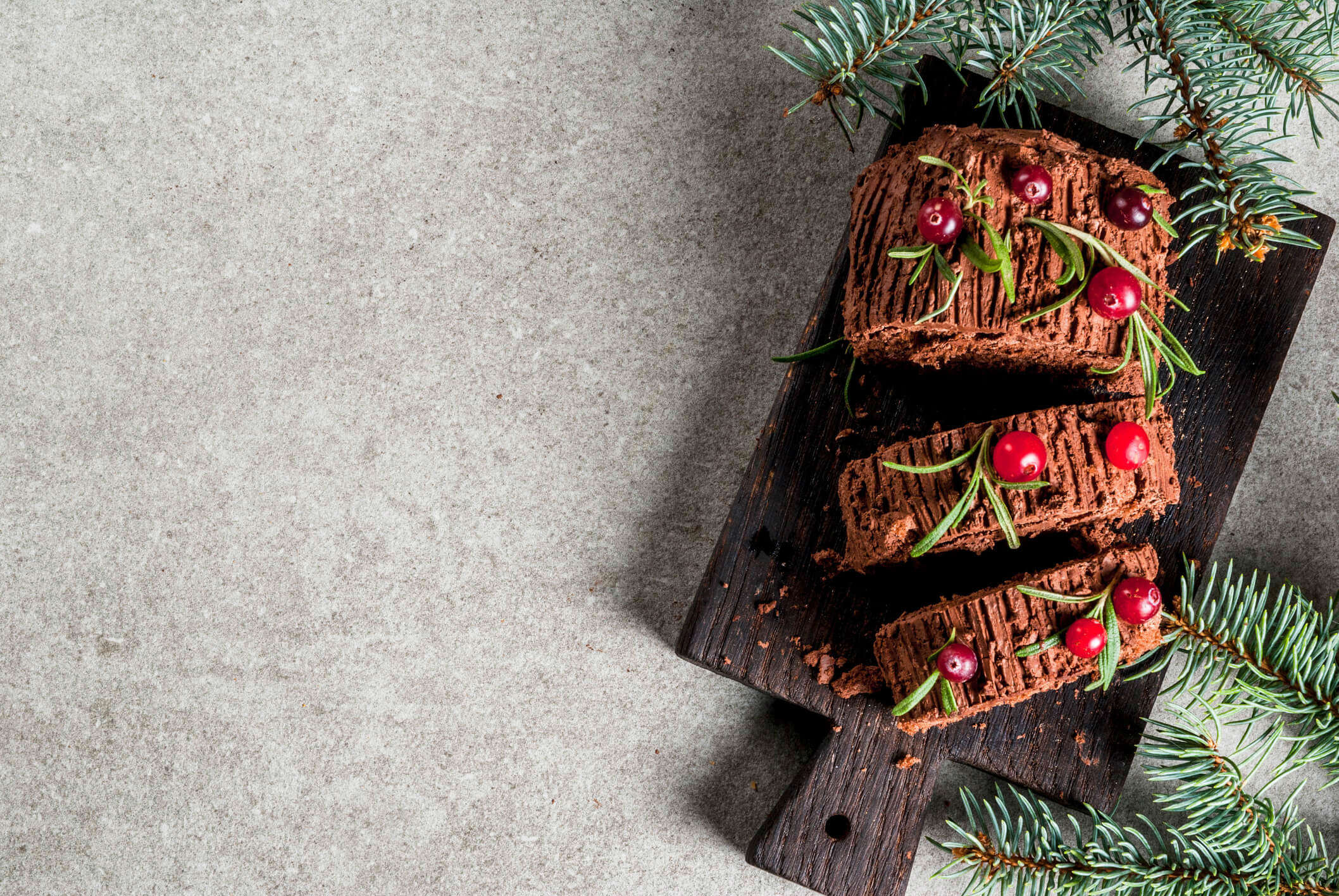 a chocolate Christmas yule log on a wooden board decorated with red cranberries and green festive herbs