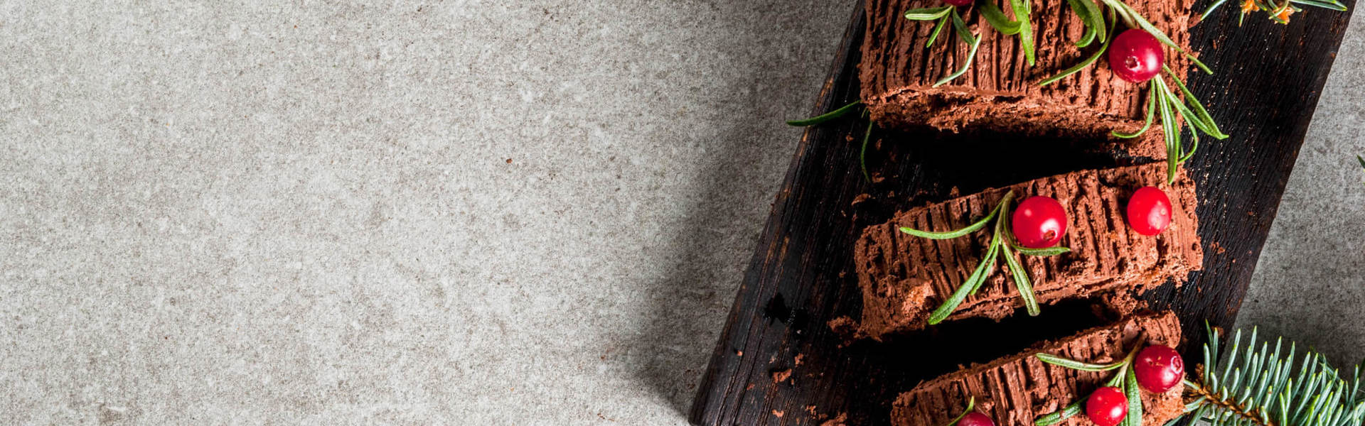 a chocolate Christmas yule log on a wooden board decorated with red cranberries and green festive herbs