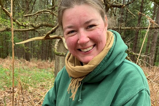 Photo of Henrietta Pringle in woodlands, smiling in a green hoodie and light green scarf.