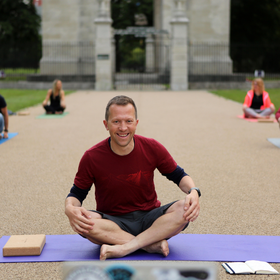 Photo of yoga teacher Dave Rennie, sat cross-legged on a yoga mat outdoors, smiling.