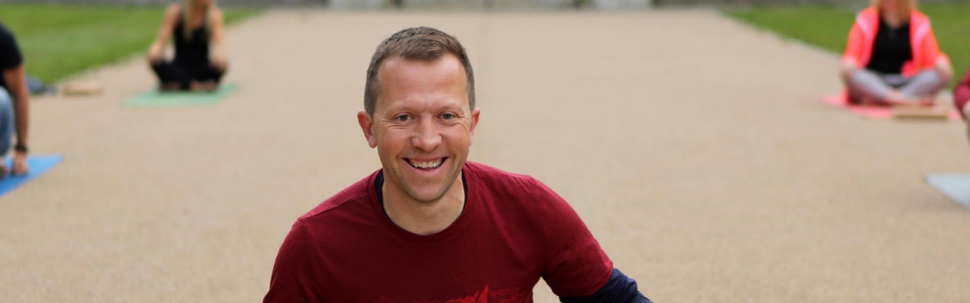 Photo of yoga teacher Dave Rennie, sat cross-legged on a yoga mat outdoors, smiling.