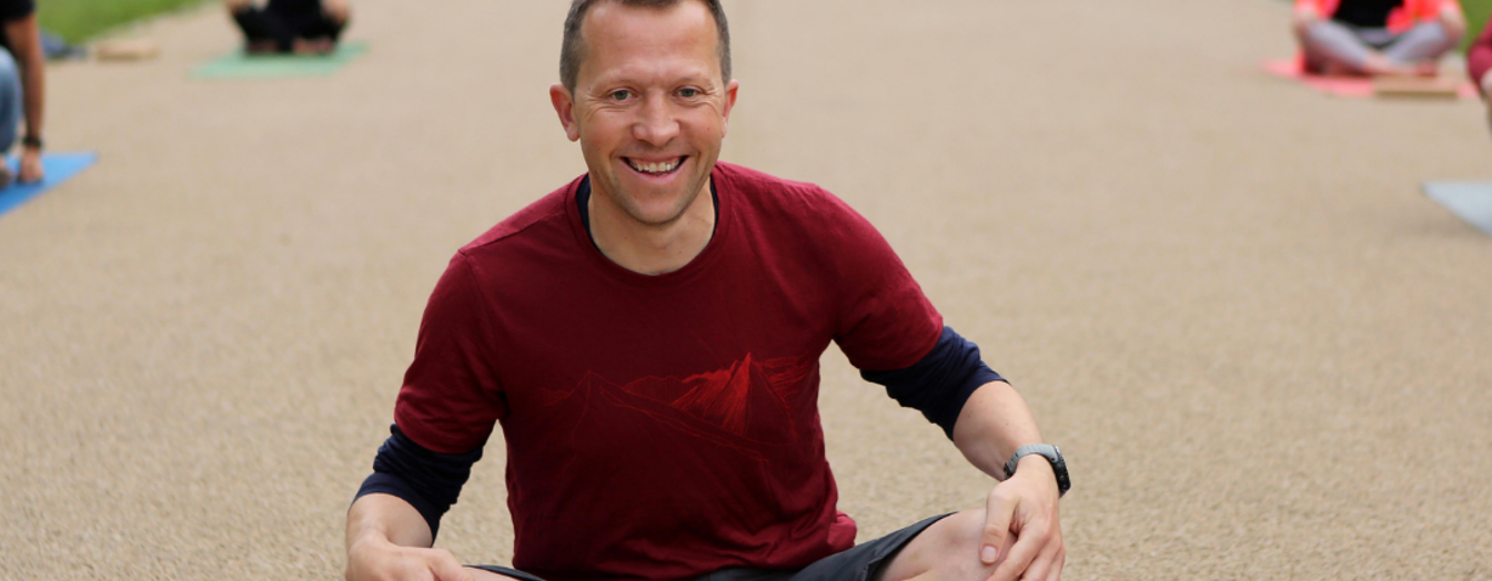 Photo of yoga teacher Dave Rennie, sat cross-legged on a yoga mat outdoors, smiling.