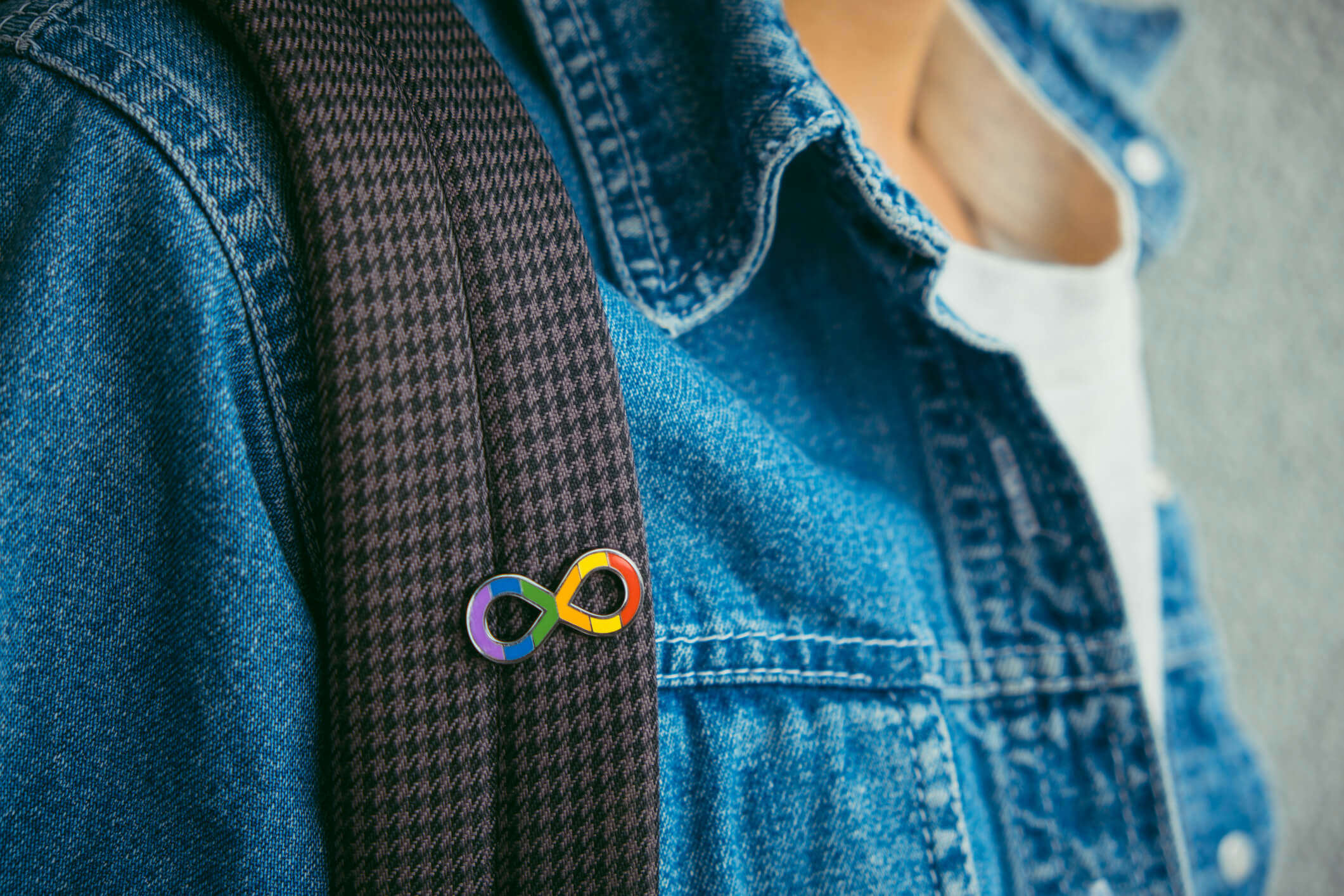 Person in a denim jacket, wearing a back pack and the rainbow infinity symbol badge, representing Autism Acceptance Month.