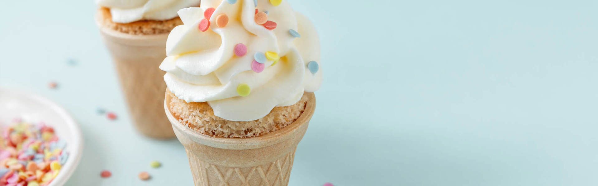 two ice cream cupcakes in ice cream cones with white icing and multicoloured sprinkles on a blue back ground