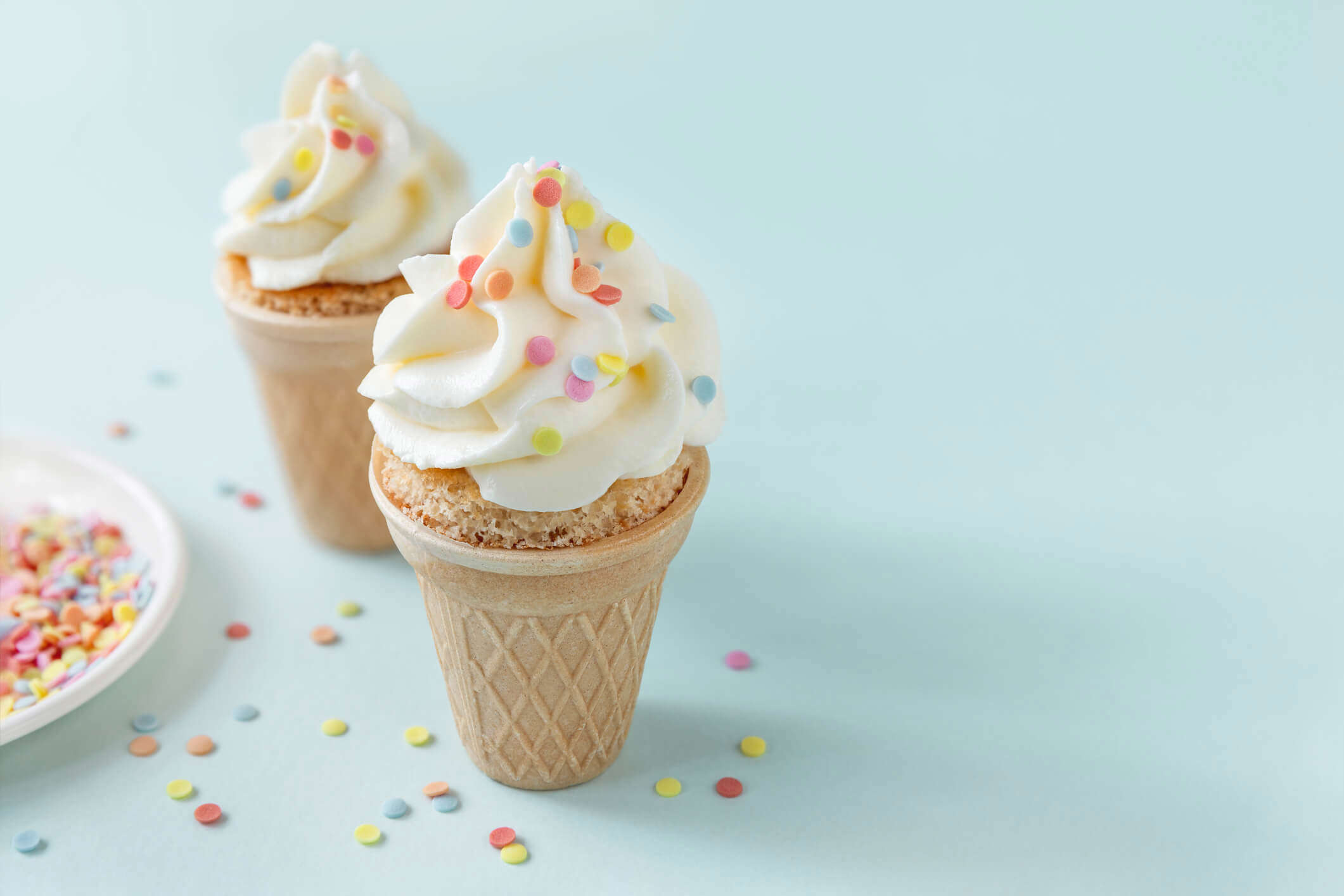 two ice cream cupcakes in ice cream cones with white icing and multicoloured sprinkles on a blue back ground